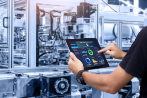 Person bedient ein Tablet mit Energie- und Produktionskennzahlen vor einer automatisierten Industrieanlage. Das Bild veranschaulicht ein Energieaudit nach DIN EN 16247 zur Identifizierung von Energieeinsparpotenzialen in der industriellen Fertigung.